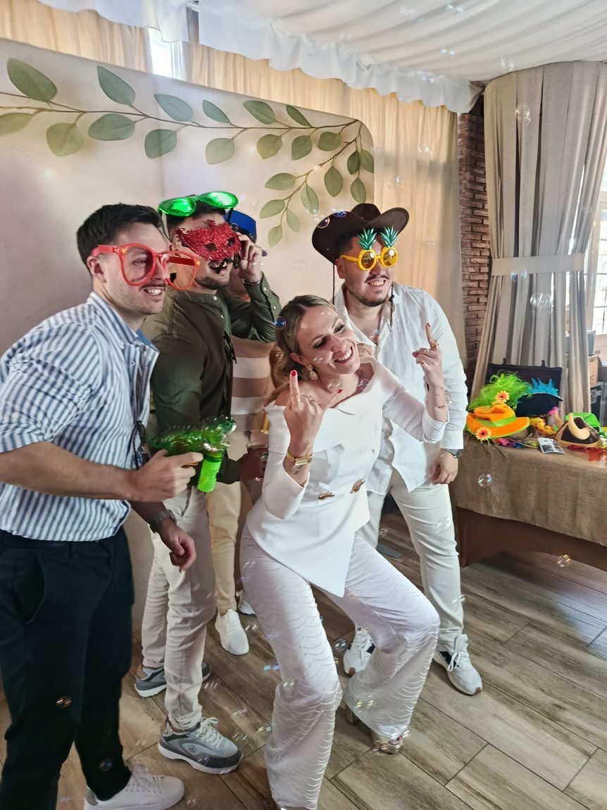 Invitados posando con gafas y sombreros de atrezzo delante del photocall de eucalipto